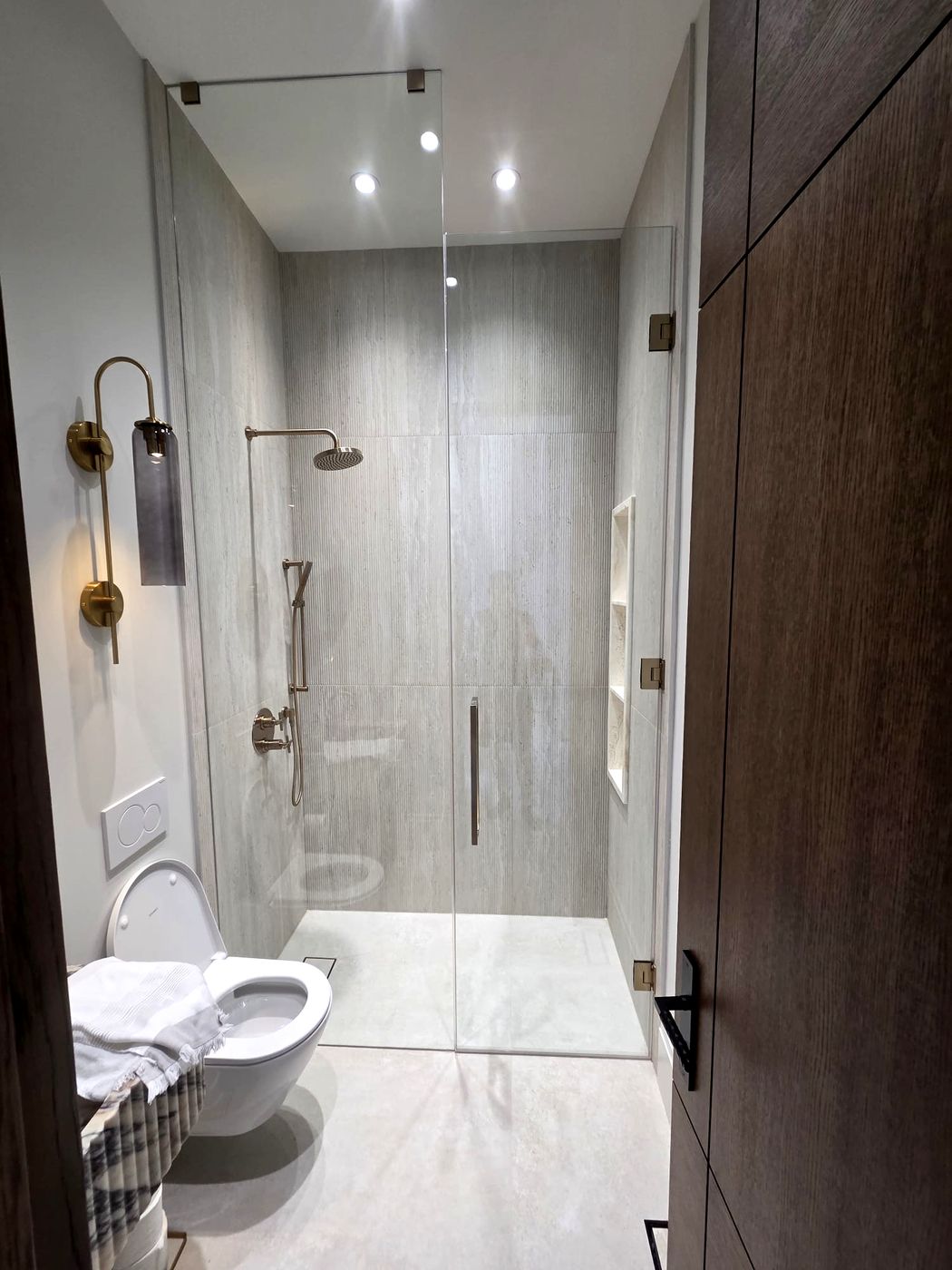 Curbless walk-in shower with frameless glass, gold rain head and hand shower, next to a wall-hung toilet