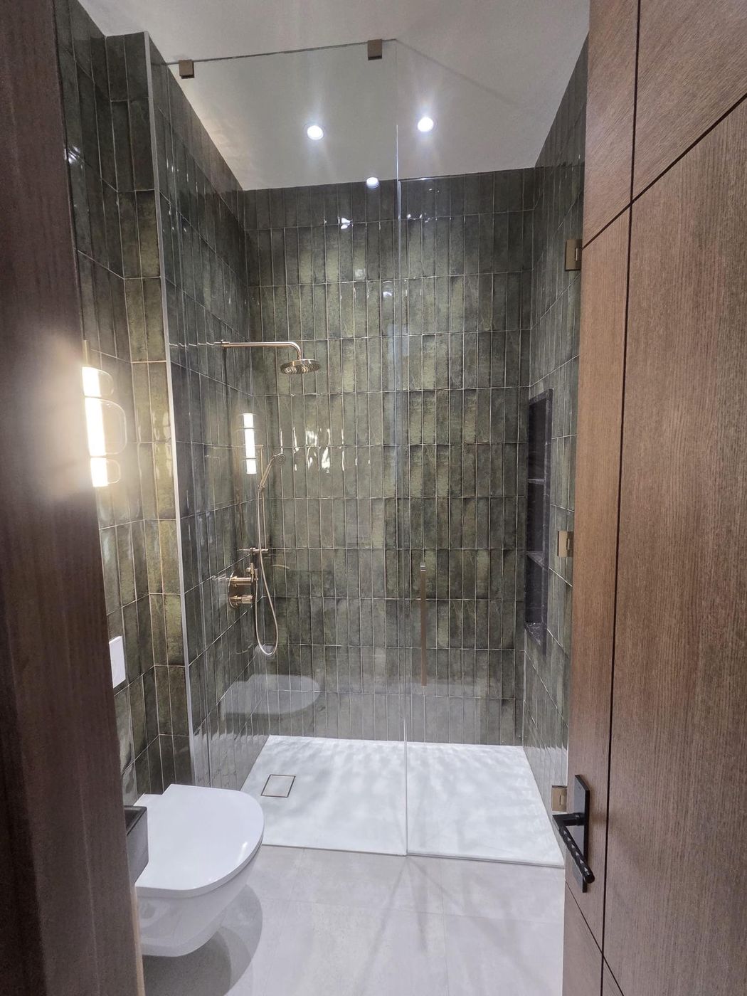 Walk-in shower with green vertical tile, brushed-gold rain head, hand shower and thermostatic valve behind a frameless glass door