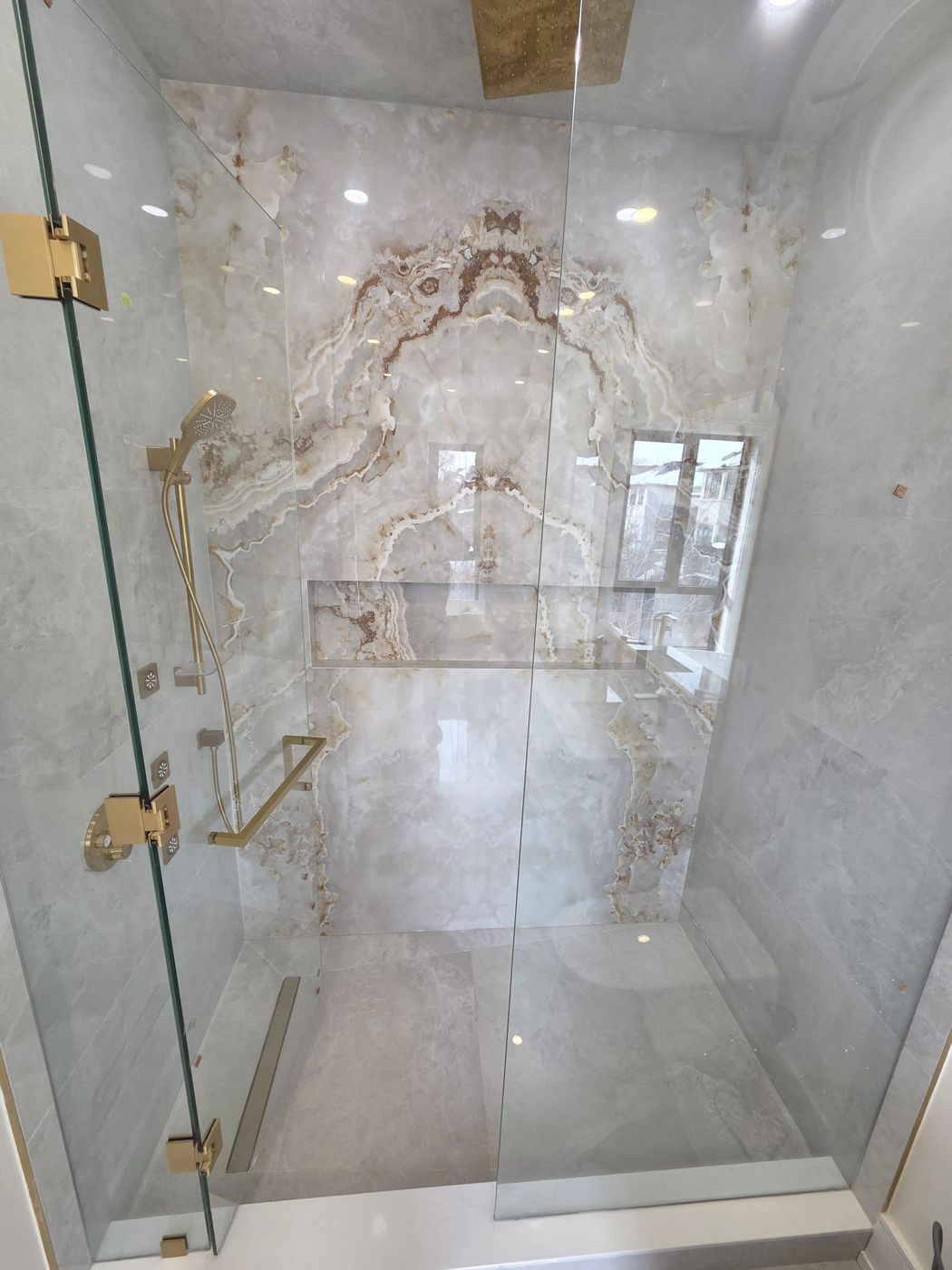 Bookmatched onyx-look slab shower with brushed-gold hand shower, body jets, towel bar and linear drain behind frameless glass